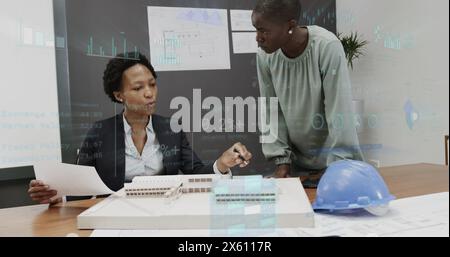 Image of graphs and changing numbers, diverse female architects discussing 3d model of building Stock Photo