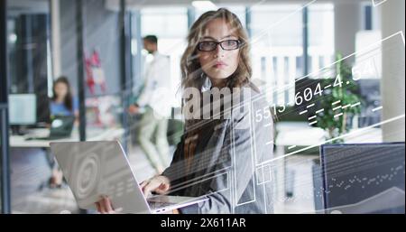 Image of graphs, loading circled and changing numbers, biracial woman using laptop in office Stock Photo