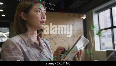 Image of multiple graphs and trading boards over asian woman scrolling on digital tablet Stock Photo