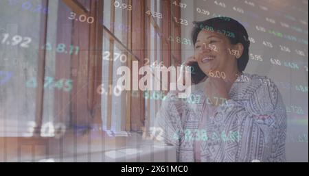 Image of trading board over biracial woman standing beside window and talking on cellphone Stock Photo
