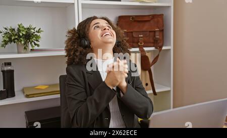 A hopeful young hispanic woman with curly hair prays in an office setting, embodying optimism. Stock Photo