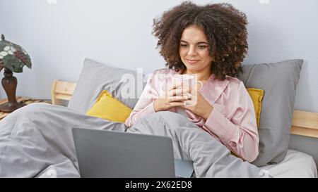 A young hispanic woman with curly hair enjoys a cozy morning in bed with a laptop and a cup of coffee, depicting modern indoor leisure. Stock Photo
