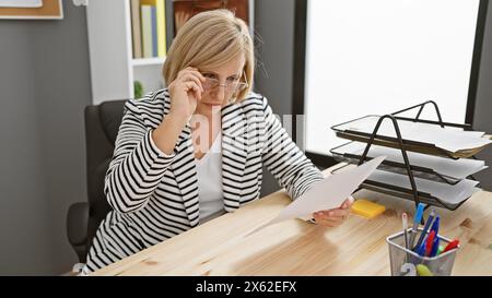 Mature blonde woman reading papers in a modern office setting, portraying professionalism and concentration. Stock Photo
