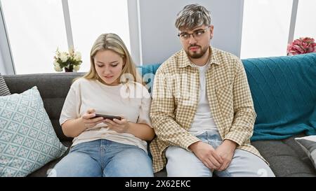 Young couple lounging on sofa with woman focused on smartphone and man looking away thoughtfully in a cozy apartment interior. Stock Photo