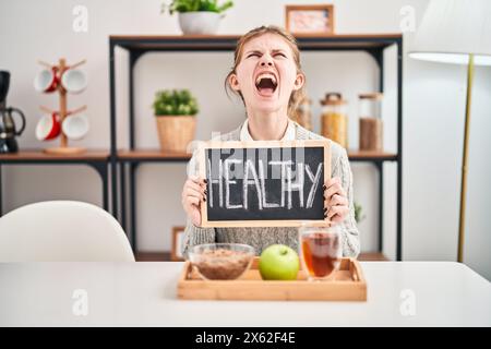 Enraged young blonde woman screaming in frustration during healthy breakfast at home, furiously holding blackboard in rage Stock Photo