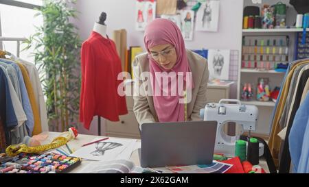 A focused woman tailor with hijab working on a laptop in a colorful atelier surrounded by garments and sewing materials Stock Photo