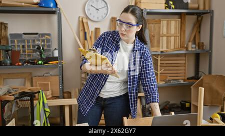 Caucasian woman with protective glasses reviews plans in a woodworking studio surrounded by tools Stock Photo