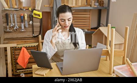 Hispanic woman multitasking in carpentry workshop with laptop and phone. Stock Photo