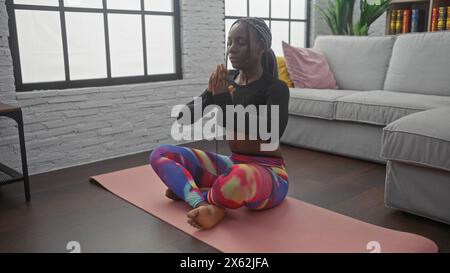 African american woman meditates on a yoga mat indoors, embodying tranquility and mindfulness in a modern home setting. Stock Photo