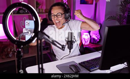 Excited woman wearing headphones in a vibrant gaming room with led lights and microphone celebrates while live streaming at night. Stock Photo