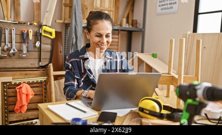 A smiling woman using a laptop in a woodworking workshop surrounded by tools and wooden pieces. Stock Photo