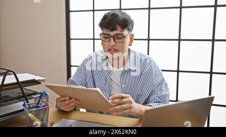 A young man in glasses works diligently in a modern office, reviewing documents beside his laptop. Stock Photo