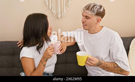 A tattooed couple shares a cozy moment with coffee on a sofa in a modern living room, portraying domestic intimacy. Stock Photo