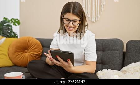 Young hispanic woman smiling while using a tablet in a cozy living room setting Stock Photo