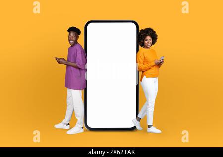Young Black Couple Standing Beside Giant Smartphone on Yellow Background Stock Photo
