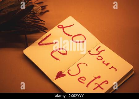 Text 'Love yourself' written in red lipstick on the sheets of open paper notebook on orange background. Self-love, healthy narcissism, motivation. Stock Photo