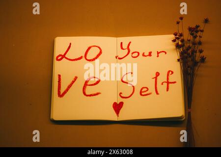 Text 'Love yourself' written in red lipstick on the sheets of open paper notebook on orange background. Self-love, healthy narcissism, motivation. Stock Photo