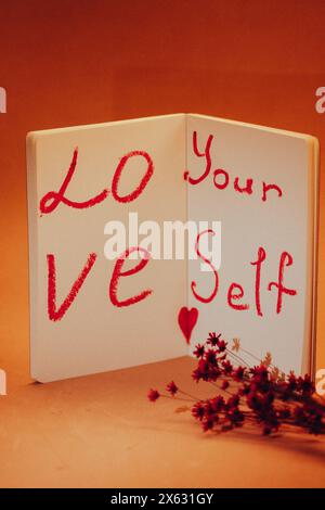 Text 'Love yourself' written in red lipstick on white sheets of open notebook Self-love, healthy narcissism concept A bouquet of small dry red flowers Stock Photo