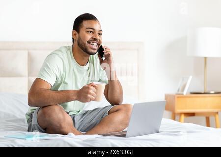 Smiling Man Sitting on Bed With Coffee, Talking on Phone Stock Photo