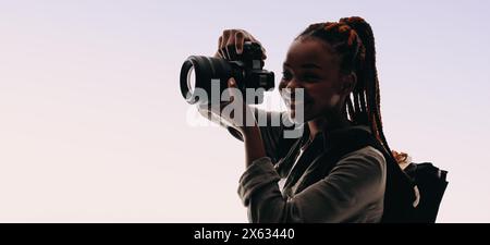 Woman with braids captures moments with her digital camera while wearing a backpack in a studio. She embodies the joy of photography, creating memorie Stock Photo