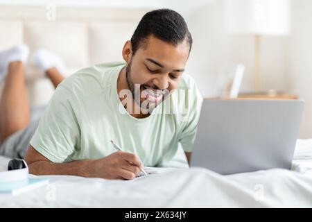 Smiling Young Man Writing Notes in Room With Laptop Open Stock Photo