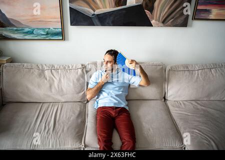 Guy coping with summer heat lying on sofa using paper fan suffocating of stuffiness, sweltering heat Stock Photo