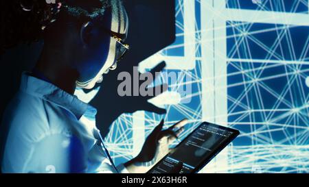 Capable supervisor overseeing dimly lit server hub, running code on tablet, doing checkup on technologically advanced machines clusters made of servers, networking systems and storage arrays Stock Photo