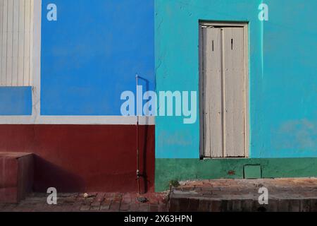 261 Colorist wall of colonial house on red-brick sidewalk, with white wood doors on an azure and electric blue, maroon, green facade. Trinidad-Cuba. Stock Photo