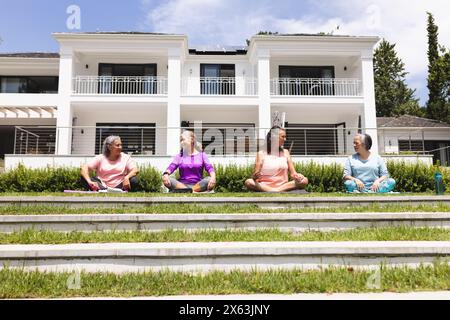 Diverse senior lady friends sit crossed-legged outdoors, basking in the sun Stock Photo