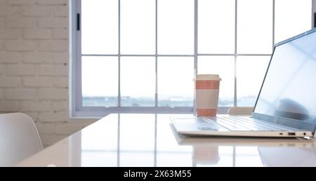 At office, laptop and coffee cup resting on white table near window Stock Photo