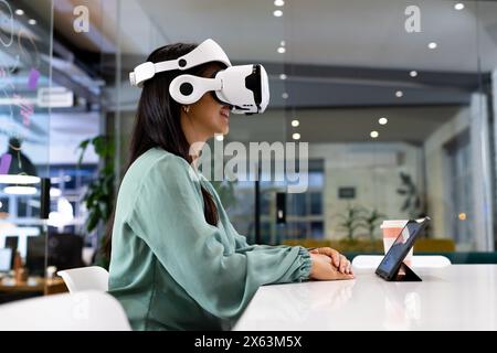At office, biracial businesswoman wearing VR headset, sitting at desk Stock Photo