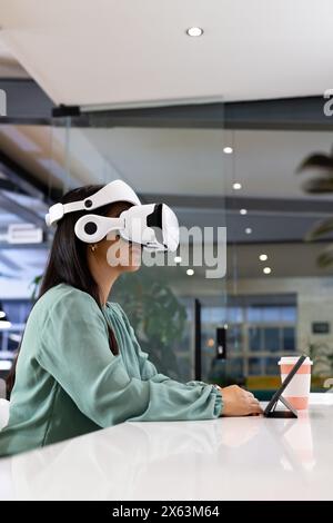 At office, biracial businesswoman wearing VR headset, sitting at desk Stock Photo