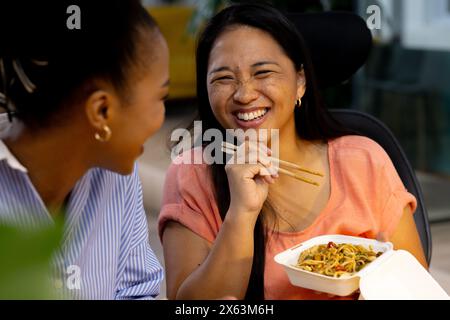 Diverse female colleagues sharing meal, laughing, and working late Stock Photo