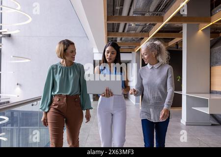 Diverse team holding laptops, walking on blue background at office Stock Photo