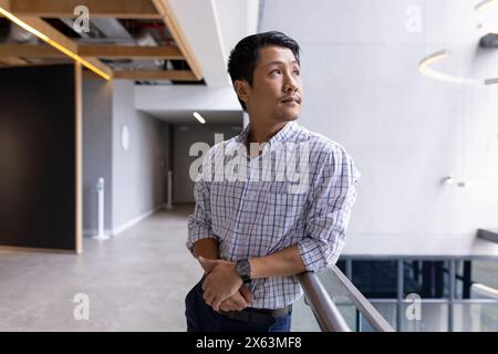 Asian male professional leaning on railing at office, looking thoughtful Stock Photo