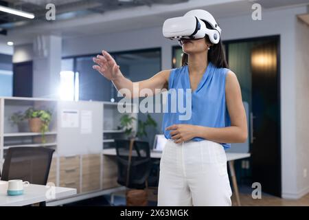 Middle-aged biracial woman wearing VR headset at office, reaching out Stock Photo