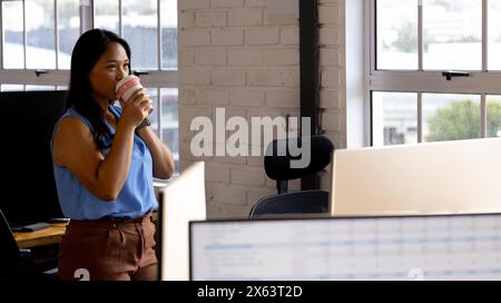 At office, biracial businesswoman drinking coffee, looking at computer Stock Photo