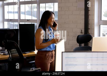 At office, biracial businesswoman holding coffee, talking on phone Stock Photo