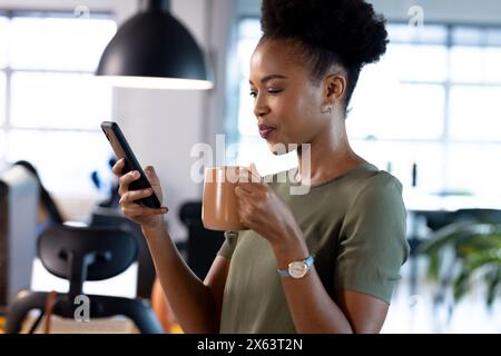 At office, African American businesswoman holding coffee, looking at smartphone Stock Photo