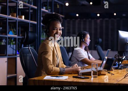 Diverse female colleagues in headsets at office, working late, focused on screens Stock Photo