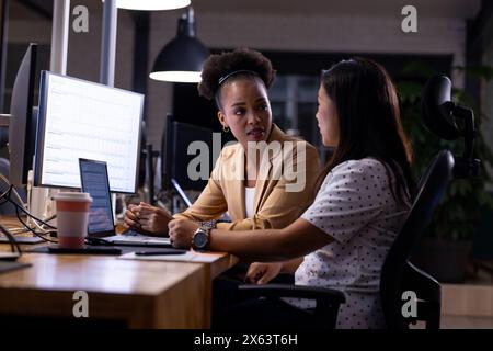 Diverse female colleagues discuss work late at office, using computers Stock Photo
