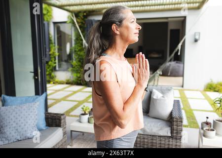 At home, mature Caucasian woman standing outside, hands together, doing yoga Stock Photo