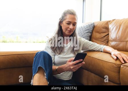 At home, mature caucasian woman sitting on sofa, looking at smartphone Stock Photo