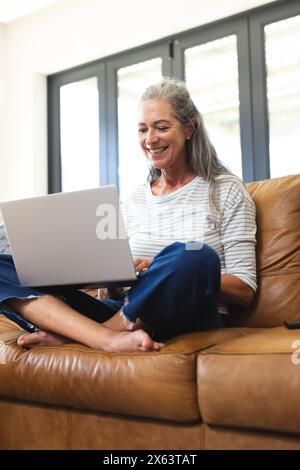At home, mature Caucasian woman sitting on a brown couch, using a laptop Stock Photo