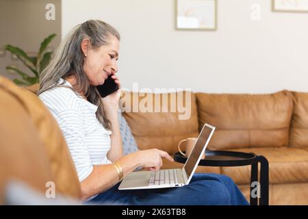 At home, mature Caucasian woman talking on phone, typing on laptop Stock Photo