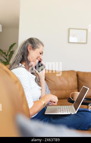 At home, Caucasian mature woman talking on phone, using laptop Stock Photo