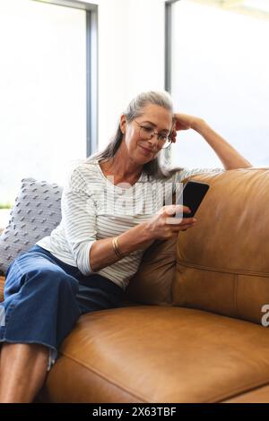 At home, mature Caucasian woman in glasses sitting on couch, looking at smartphone Stock Photo