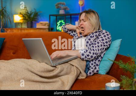 Bored sleepy young Caucasian young woman working on laptop computer, yawning, leaning on hand sitting on sofa with blanket. Exhausted tired freelancer workaholic girl sleeping in living room at home. Stock Photo