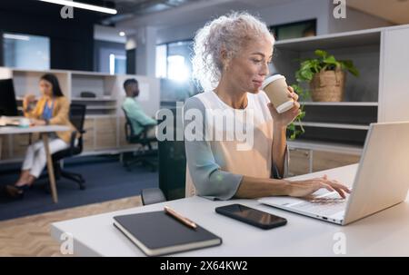At office, diverse team with Caucasian woman drinking coffee, using laptop Stock Photo