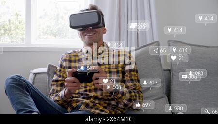 Image of social media icons over caucasian man wearing vr headset playing games at home Stock Photo